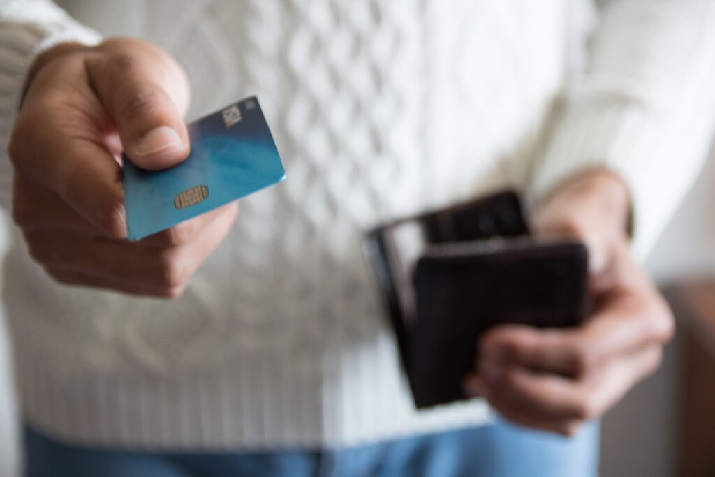 A photo showing a man in a white sweater holding a wallet in one hand and holding out his credit card with another.