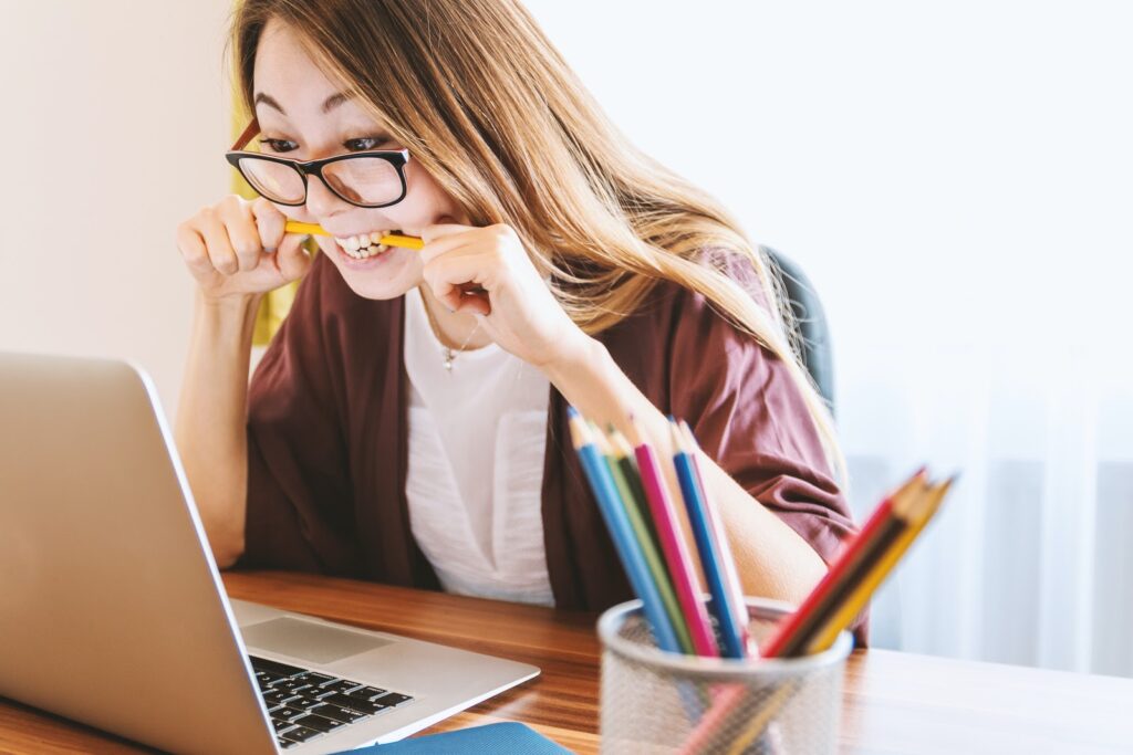 A woman sitting at her laptop, looking angrily at the screen and biting a pen in frustration.